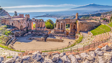 Ruins of the ancient greek theater of Taormina, Sicily the Etna with its double smoke tail in the background above the morning sun lit Giardini-Naxos bay of the Ionian see.