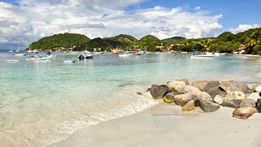 Pale aqua water washes along a white sand beach, overlooked by green hills.