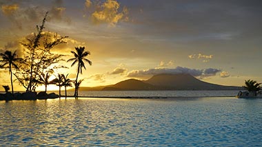 Sun setting behind a few palms and far off mountain island surrounded by water.