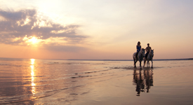 A couple rides horses on the beach at sunset.