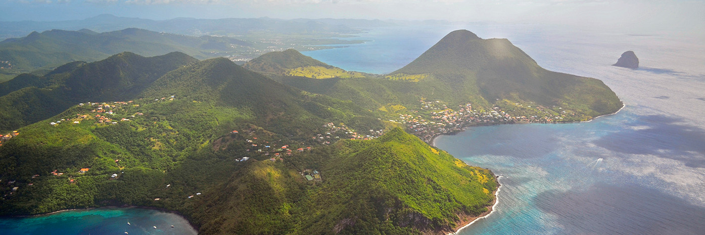 In an aerial view of an island, mist hangs over the lush greenery and blue water.