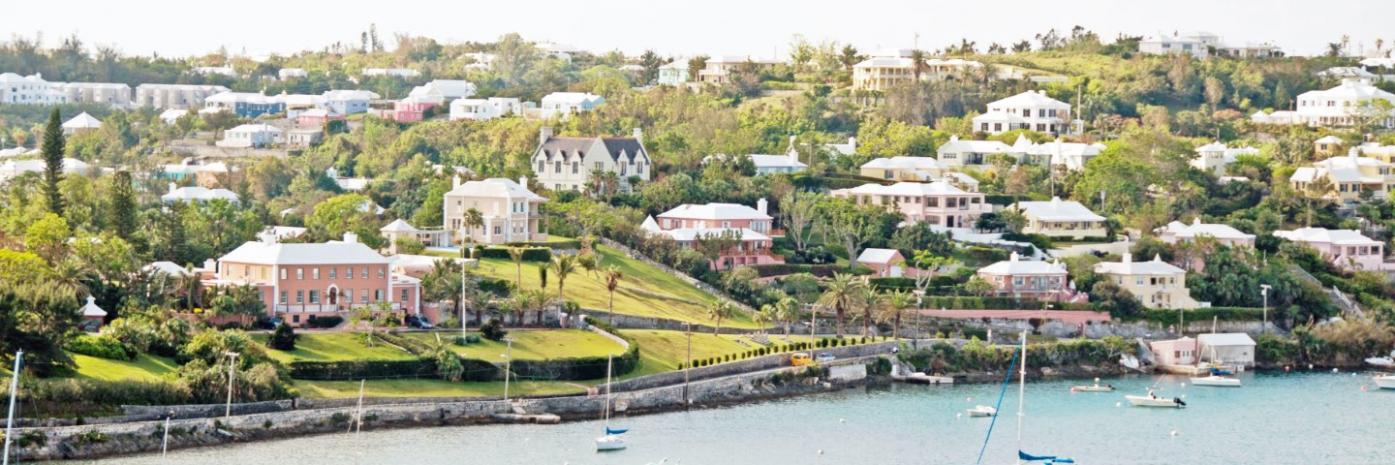Pink and white hued houses line a hill in front of the water, dotted with boats.
