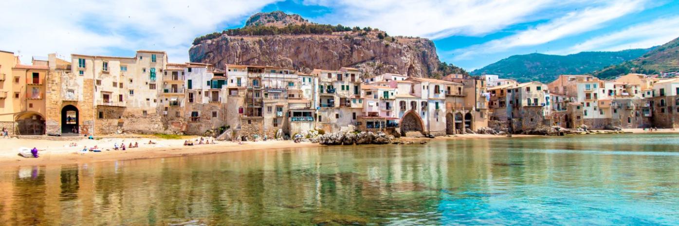 Blue green water before a sandy shore with old buildings.