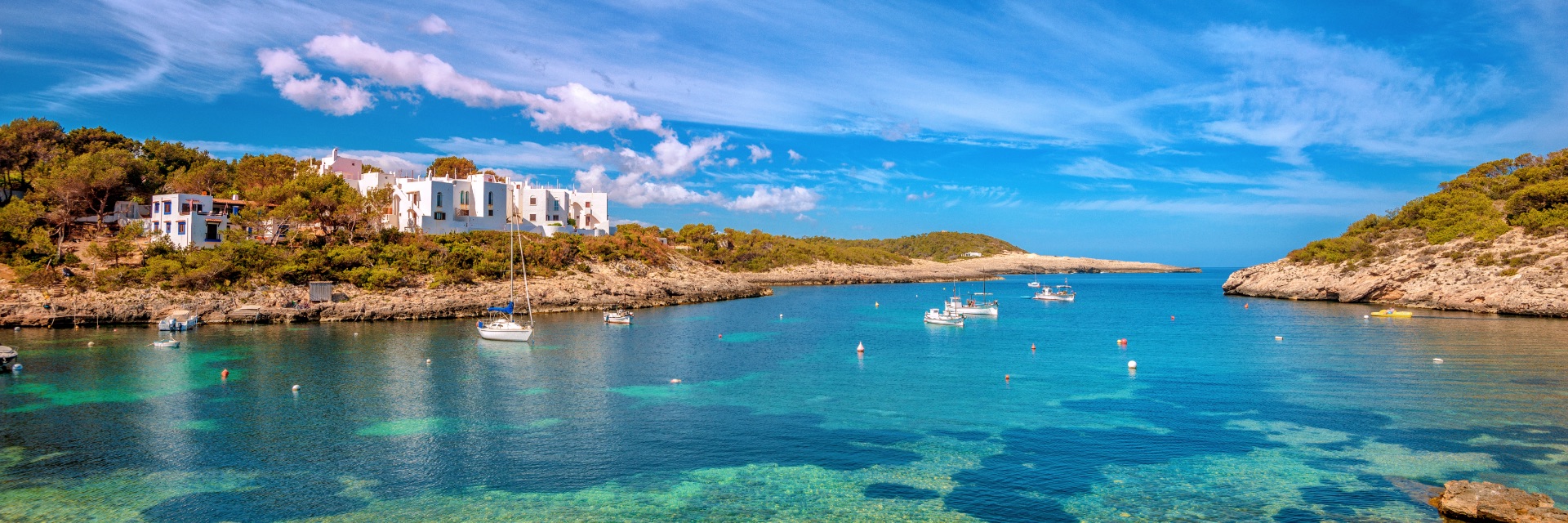 Sailboats scattered in the bright blue harbor looking inland to orange and lush green cliffs.