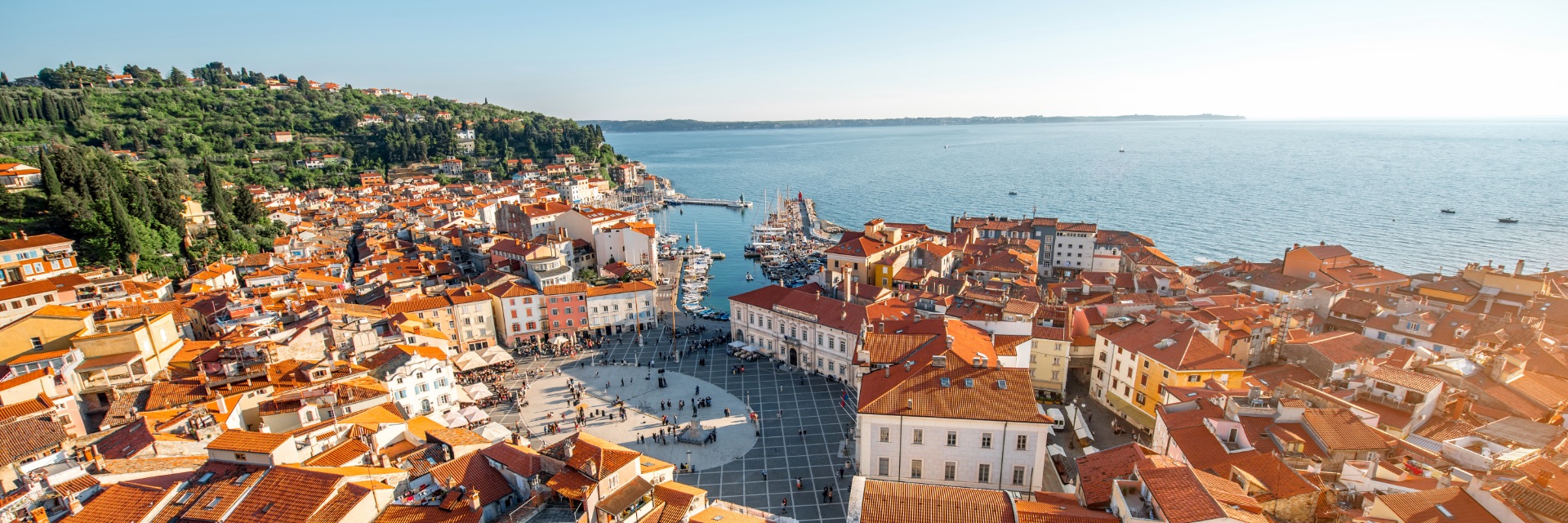 The aerial view of an old town, featuring European buildings with terracotta roofs, a harbor and lush green hills in the background.