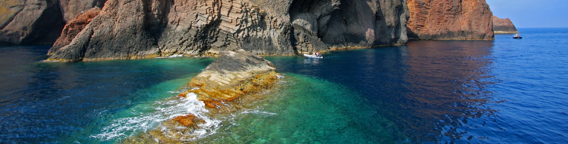Red and brown cliff surrounded by blue waters with boats sailing nearby.