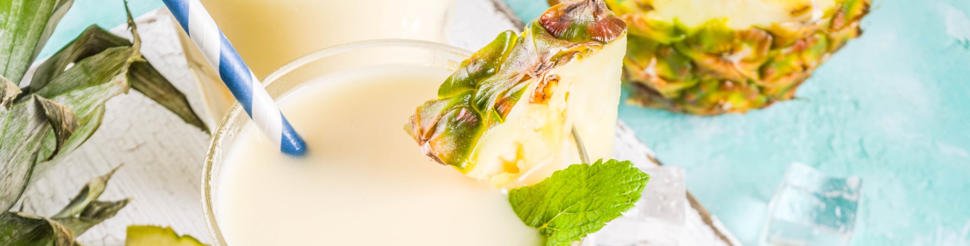 Two drink glasses of piña colada on a wooden board surrounded by open coconuts and pineapple slices.