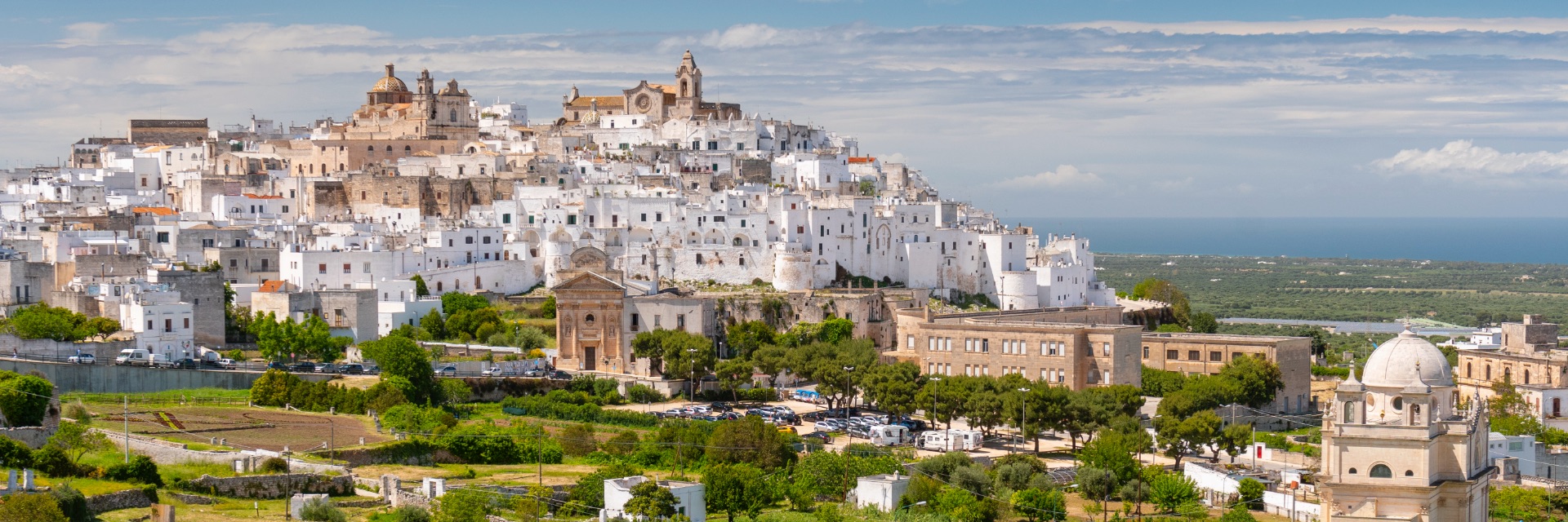 White and tan buildings in a city on a hill with clouds over blue sea behind.