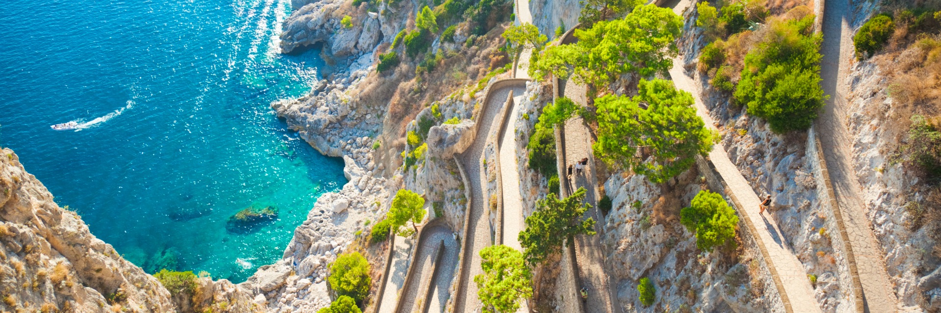 An aerial view of winding roads on a cliff overlooking bright blue-green water.