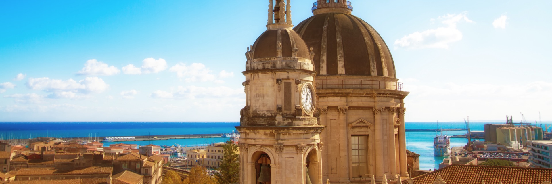 A clock tower among tan buildings in the sun, in front of bright blue water.