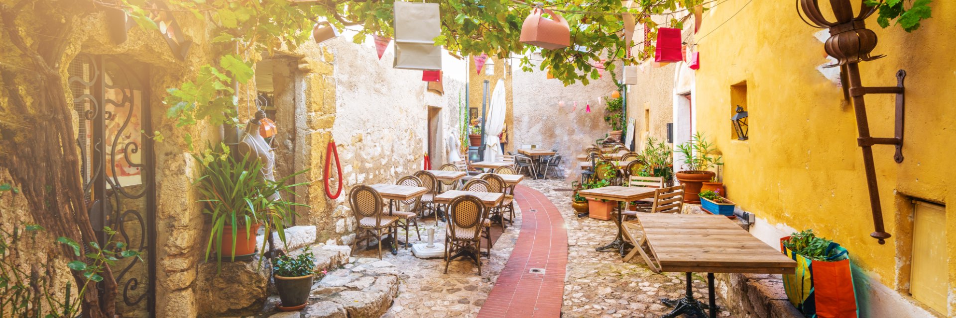 A small cobblestoned street lined with cafe chairs and green leaves.