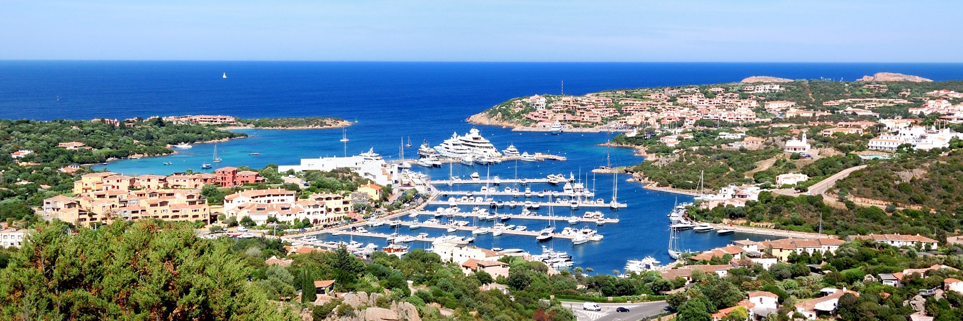 A marina with boats docked in blue water, surrounded by green land dotted with buildings.