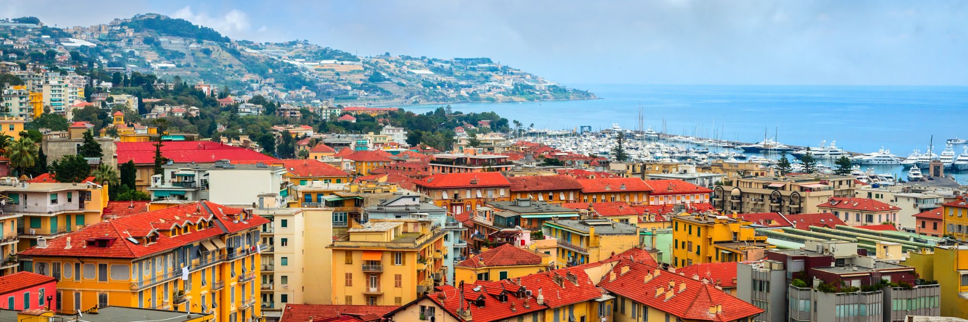 Yellow buildings with orange roofs in a city along the water, with mountains in the background.