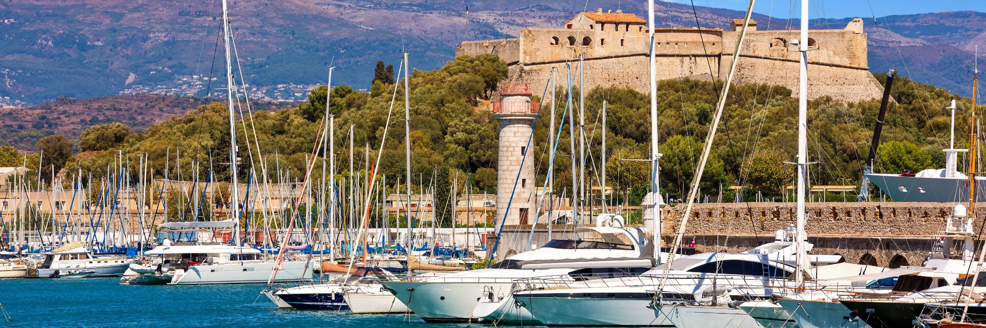 Several sailboats docked in a harbor with a stone fortress in the background.