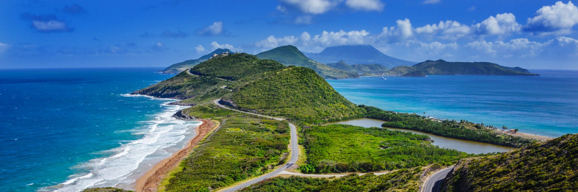 A road winds down the lush green hills of St. Kitts island, with bright blue water on either side.