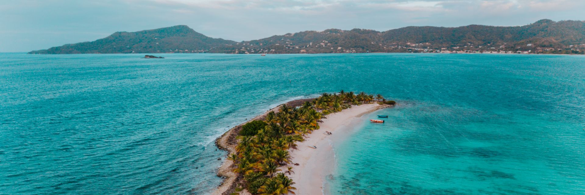 The aerial view of a peninsula of sand and palm trees extending into bright blue waters with a large island in the background.