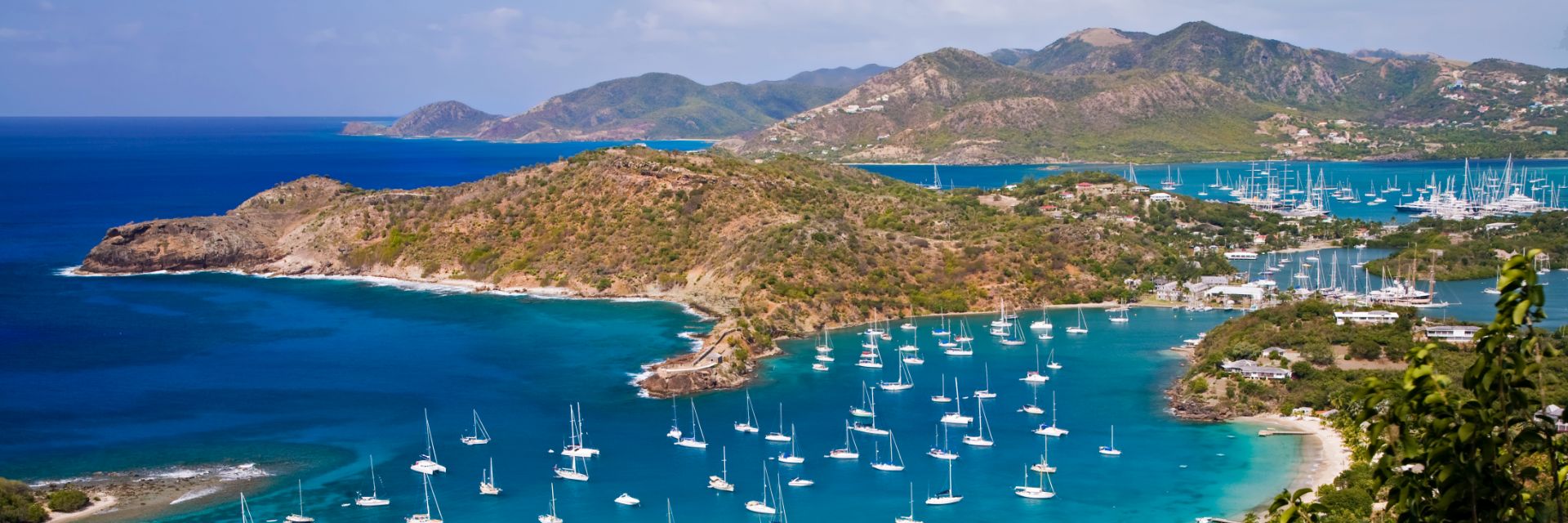 An aerial view of Caribbean islands Antigua and Barbuda with harbors and coves dotted with white sail boats in the foreground.