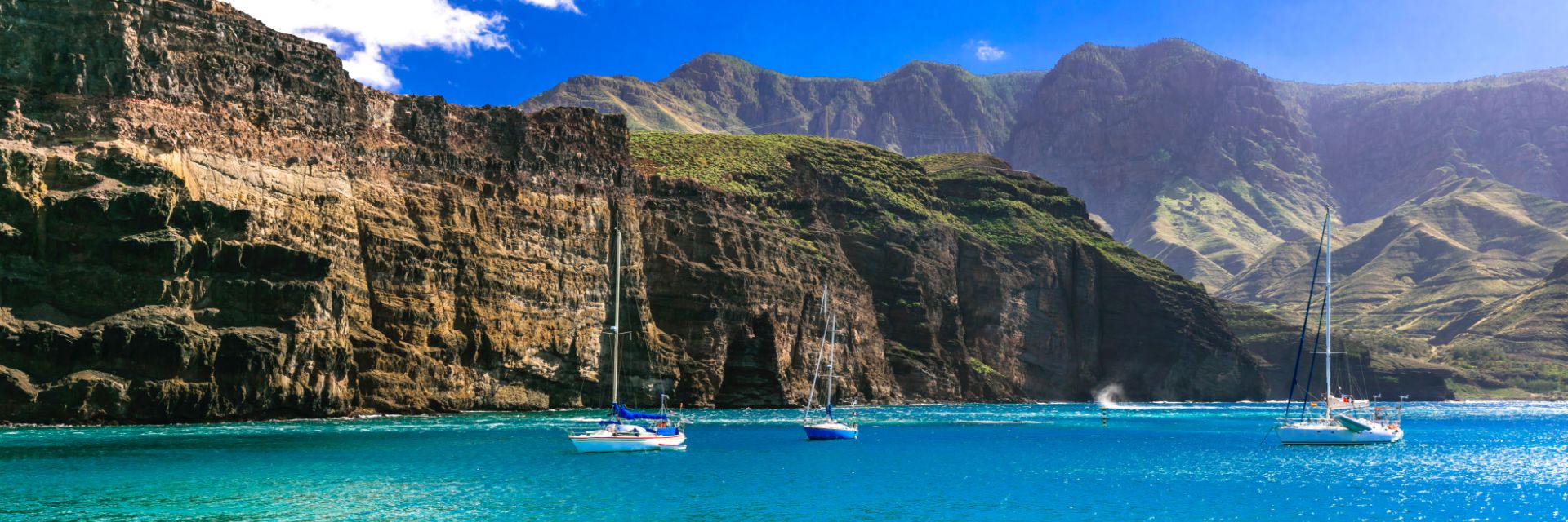 Giant cliffs tower over the bright blue sea and three sailboats at anchor off the coast of Gran Canaria, Canary Islands.