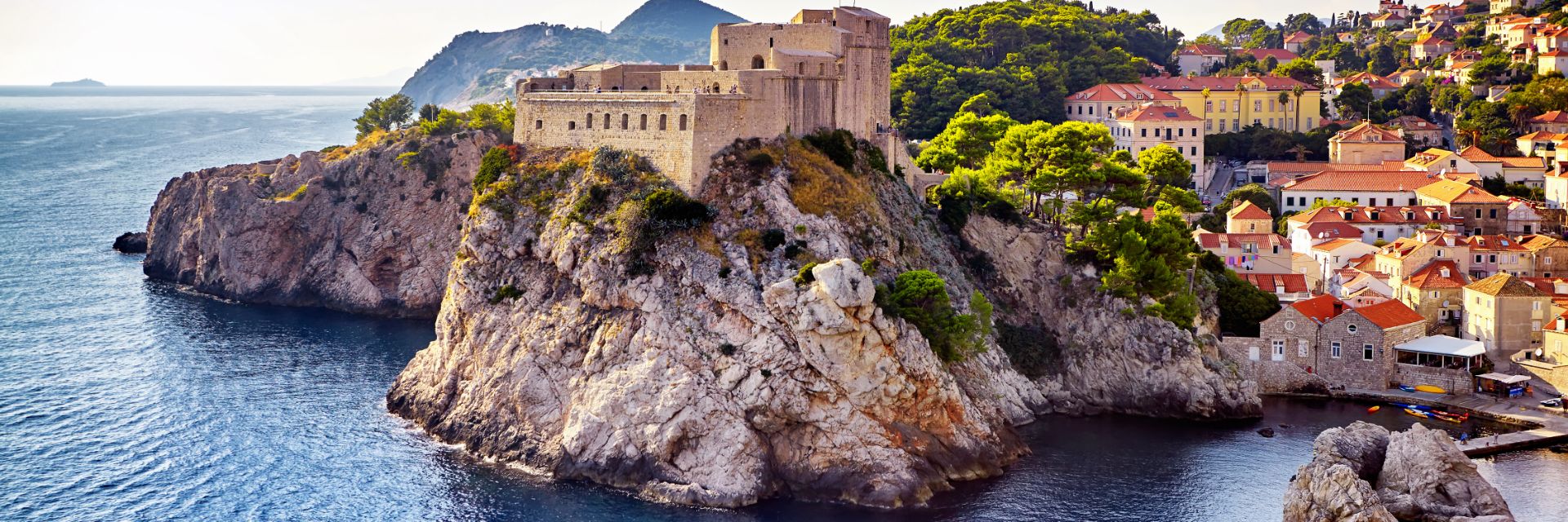 A medieval fort on top of a cliff overlooking the sea with the orange-roofed buildings of Dubrovnik, Croatia, in the background.