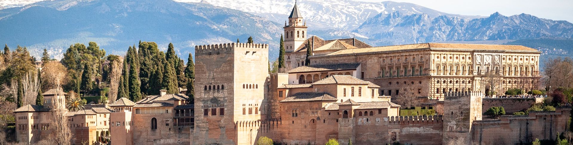 The view of the Alhambra palace surrounded by trees with snowcapped mountains in the background.