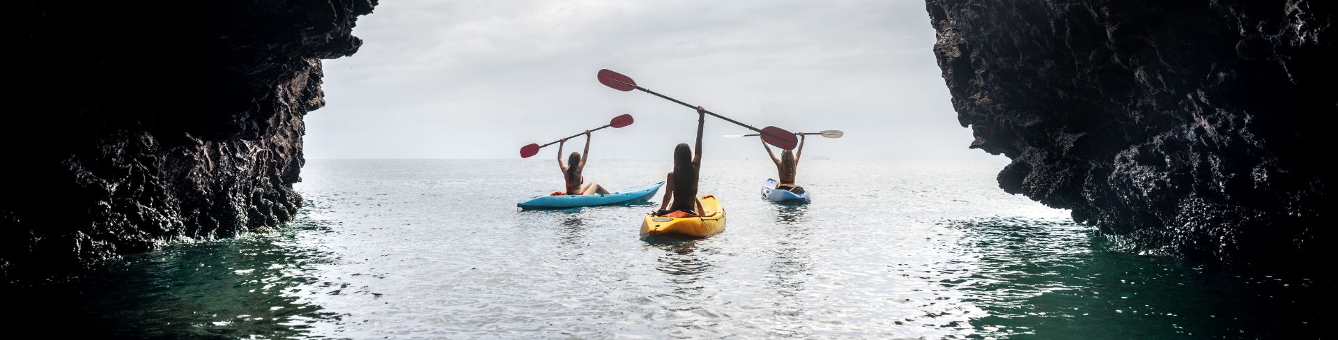 Three women facing away from the camera in kayaks, holding their paddles above their heads.