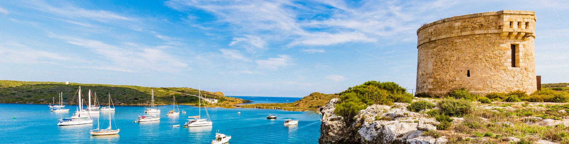 La Mola fortress sits on a cliff overlooking a small blue bay dotted with sail boats at anchor.