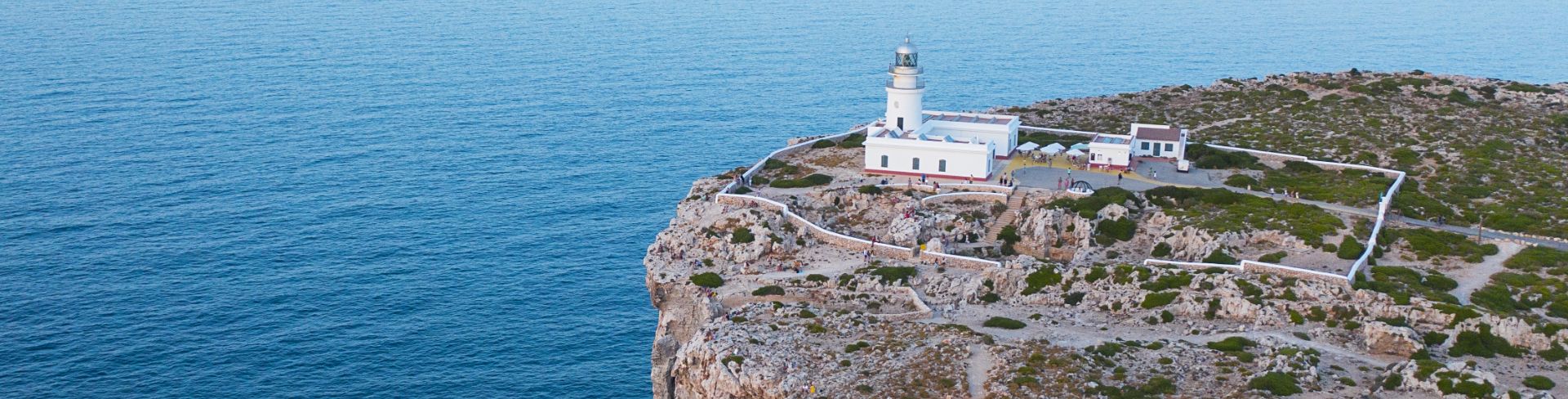 An aerial view of the top of a rocky cliff featuring a whitewashed lighthouse overlooking the blue ocean.