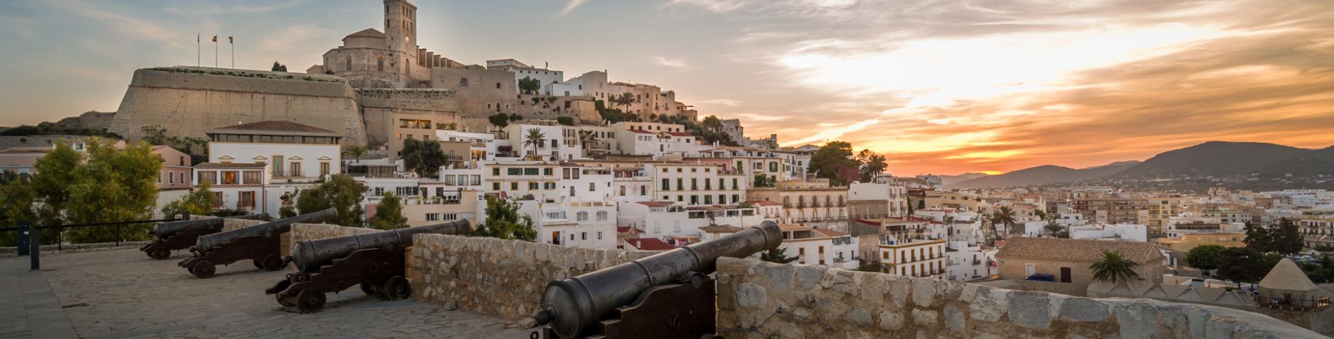 Old cannons positioned in a square overlooking the white buildings of Ibiza at the foot of a fortress.