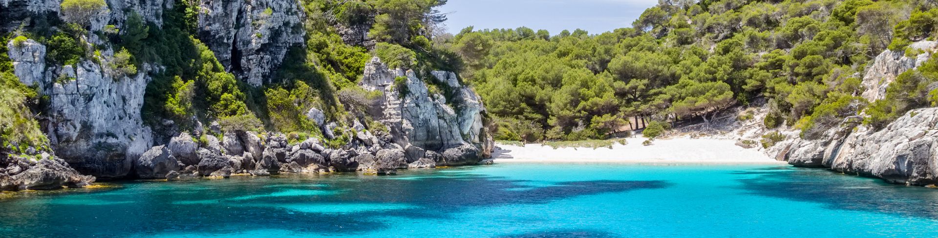A view of a white sand beach from turquoise waters, surrounded by limestone rocks covered in greenery.