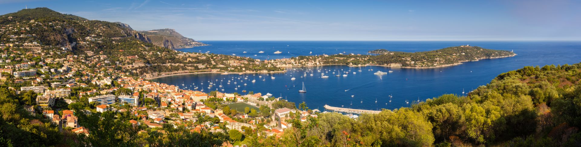 A panoramic view of a rolling green hills, and old French town and bay dotted with yachts and sail boats.