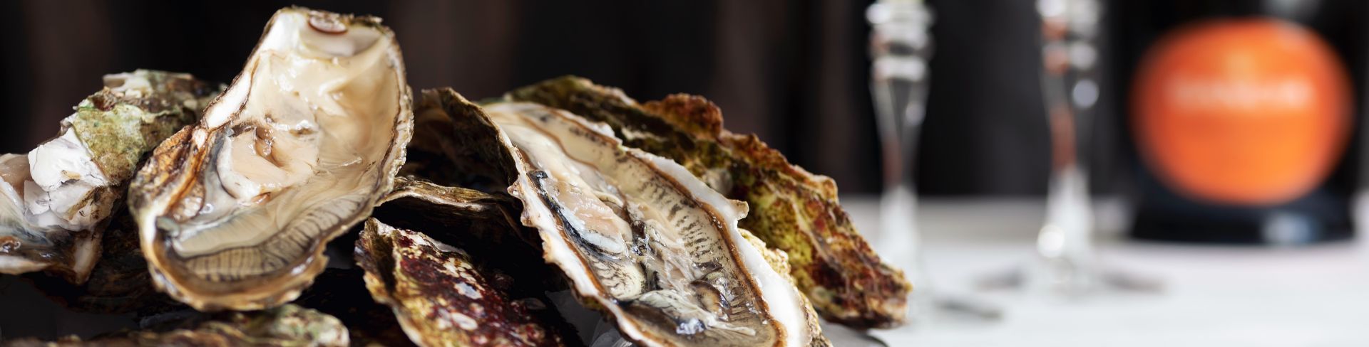 A pile of shucked oysters in the half shell on a table with two flutes of sparkling wine in the background.