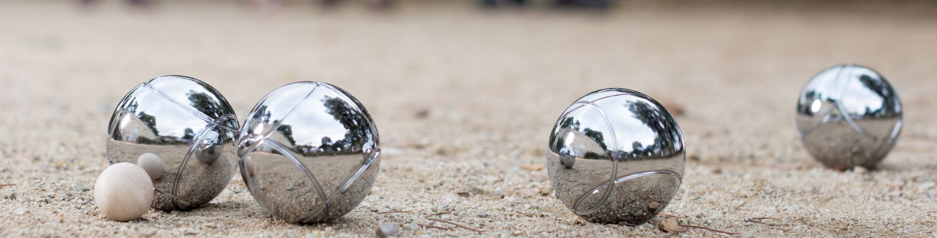Four silver pétanque balls rest on the dirt near the target ball with players in the background.