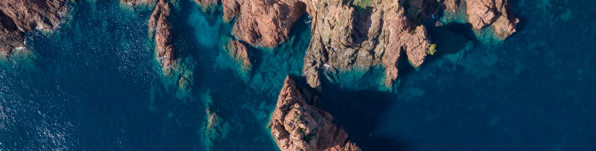 Top-down aerial view of a rocky coastline featuring reddish cliffs rising out of the blue-green sea.