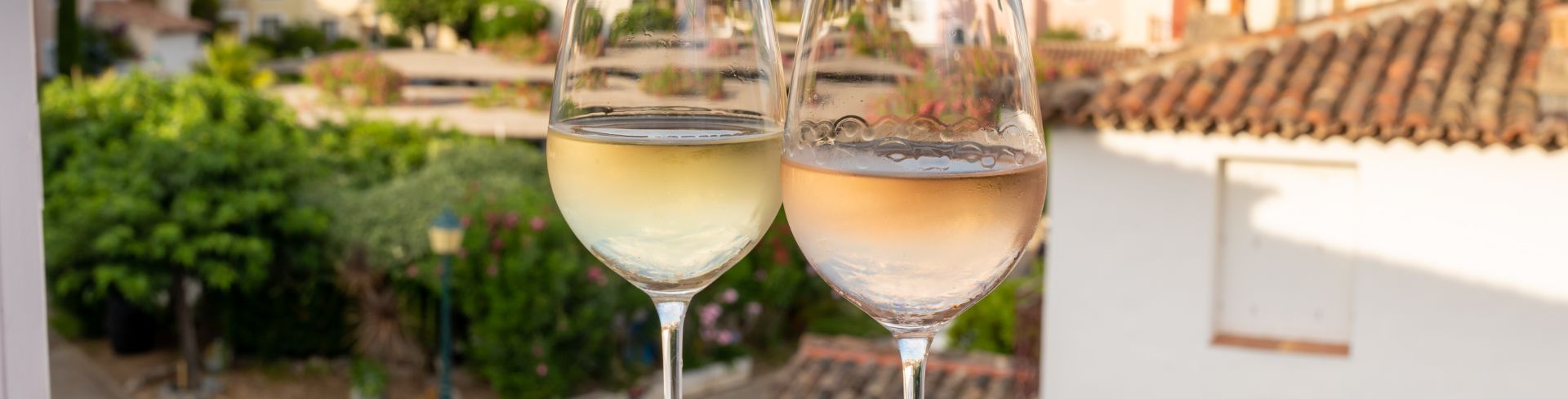 A glass of white wine and rose wine sit on a table overlooking a French Riviera town of whitewashed buildings.