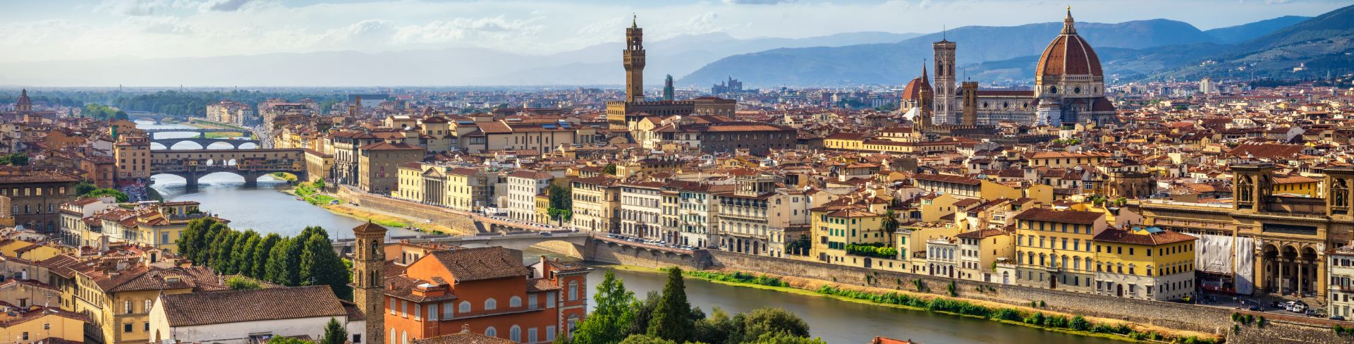 An aerial view of the city of Florence with bridges crossing over the river and orange, yellow and tan buildings on both sides.