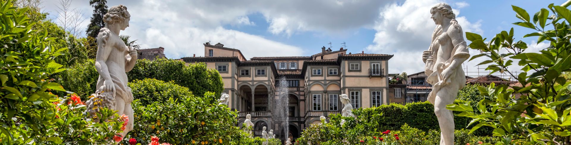 A pathway through a lush garden full of roses and statues with a fountain and villa the end of the path.