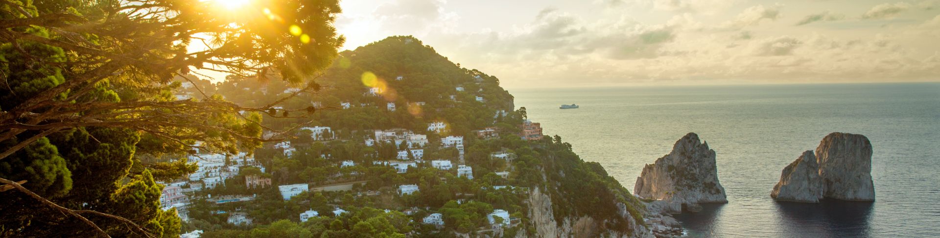 A panoramic view of the homes of Capri overlooking the water and Faraglioni rock formations at sunrise. 