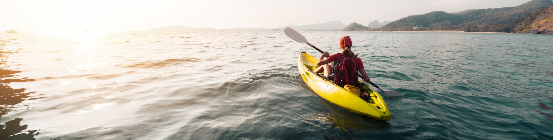 The back of a woman in a yellow kayak paddling on the sea with a hilly coastline to the right.