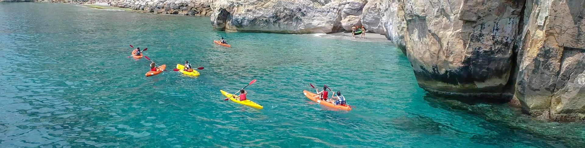 Six kayakers paddling on blue-green water alongside gray cliffs and remains of ancient villas.