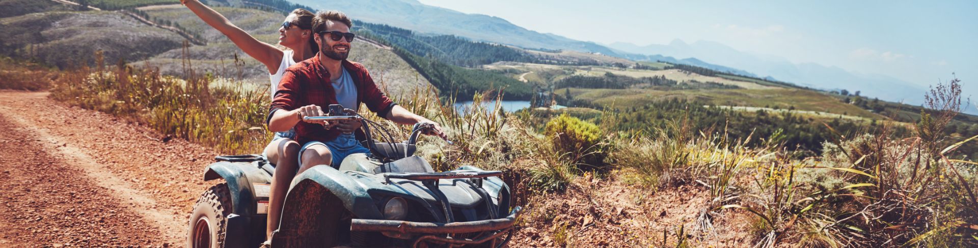 A couple riding a quad on a red gravel trail on a hilltop overlooking rolling hills.