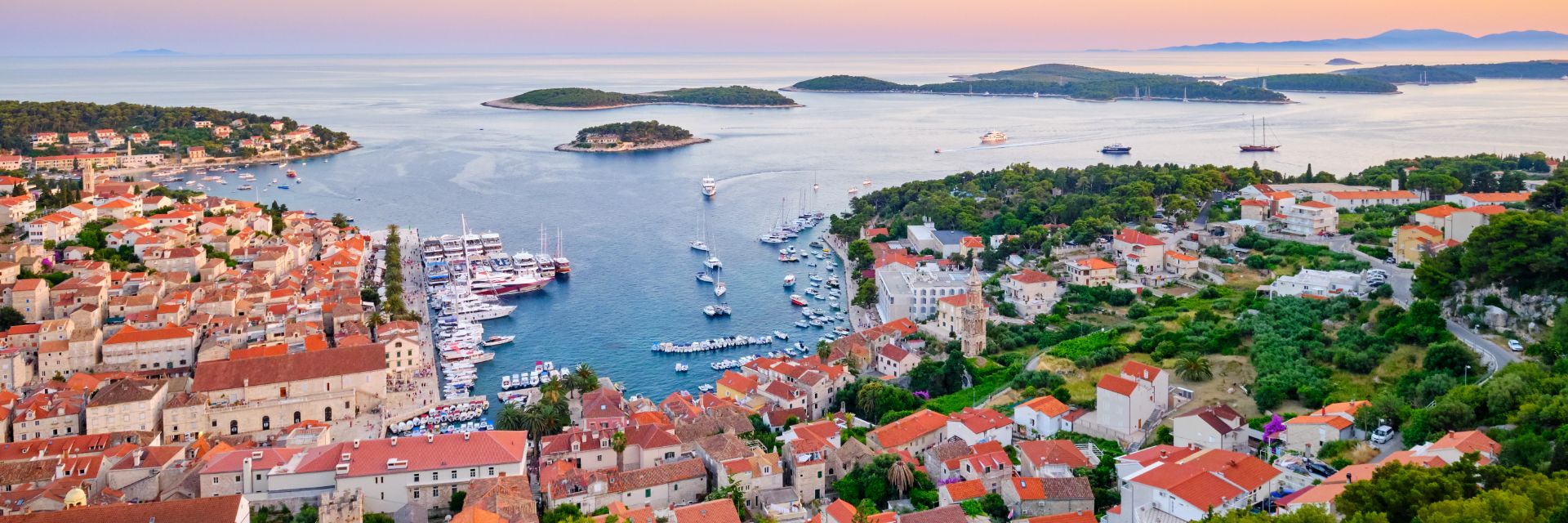 The aerial view of a harbor at sunset, encircled by a medieval town of orange roof buildings.