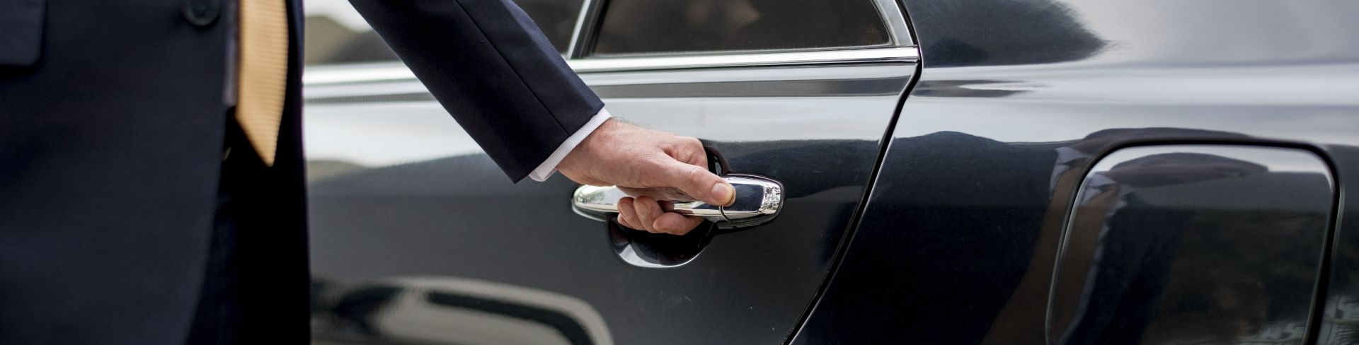 A man in a suit is poised to open a town car door with his hand on the handle.