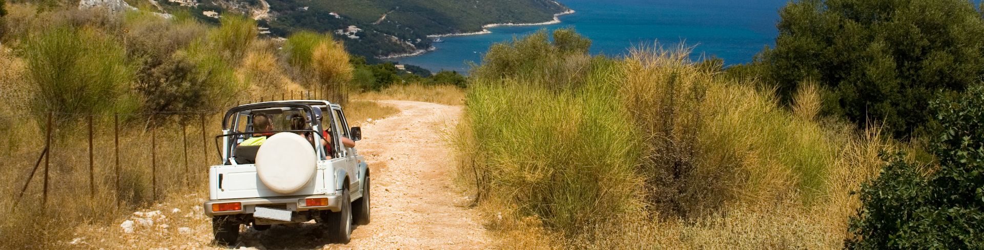 A white jeep drives away on a gravel road with the greenery of the island and blue sea in the background.