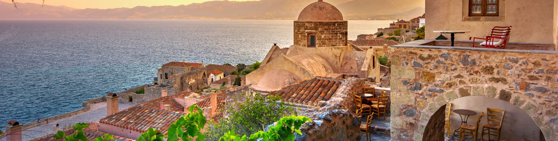 Old stone house on the sea at sunset with mountains in the distance.