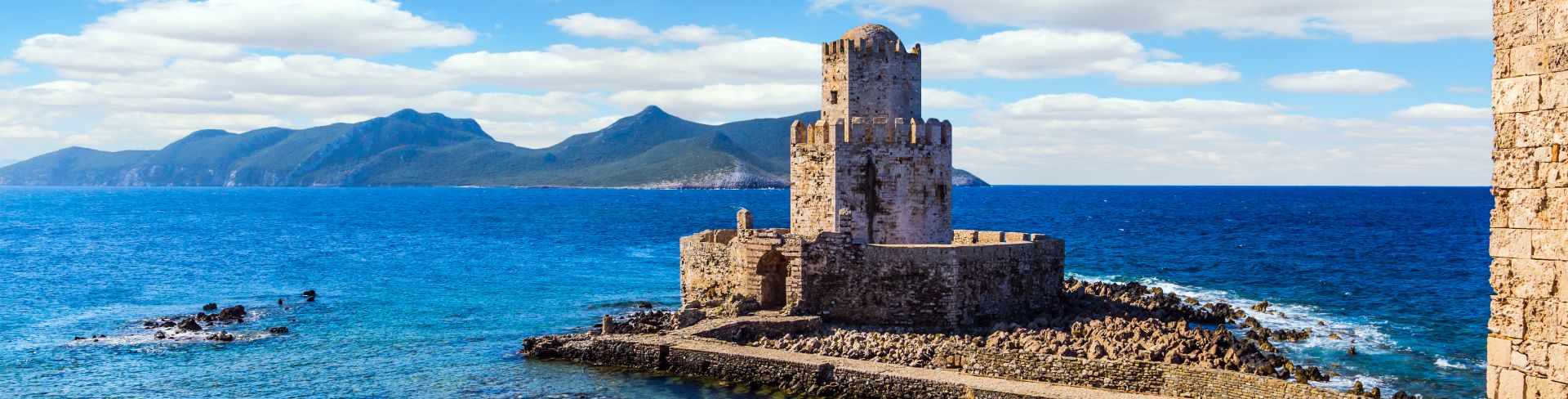 A three-tiered watchtower sits on a rocky peninsula overlooking the blue sea with green mountains in the background. 