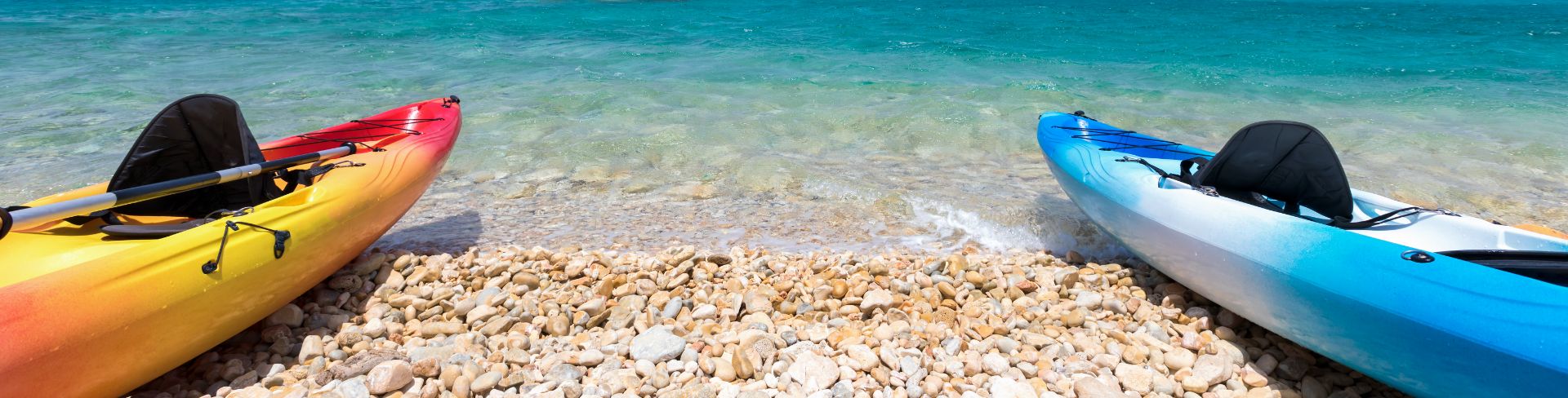 An orange and a blue kayak sit side by side on the edge of a rocky beach and clear blue waters.