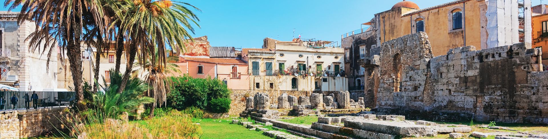 Ruins of Temple Apollo in Siracusa, Italy, featuring a grassy area with palm trees.