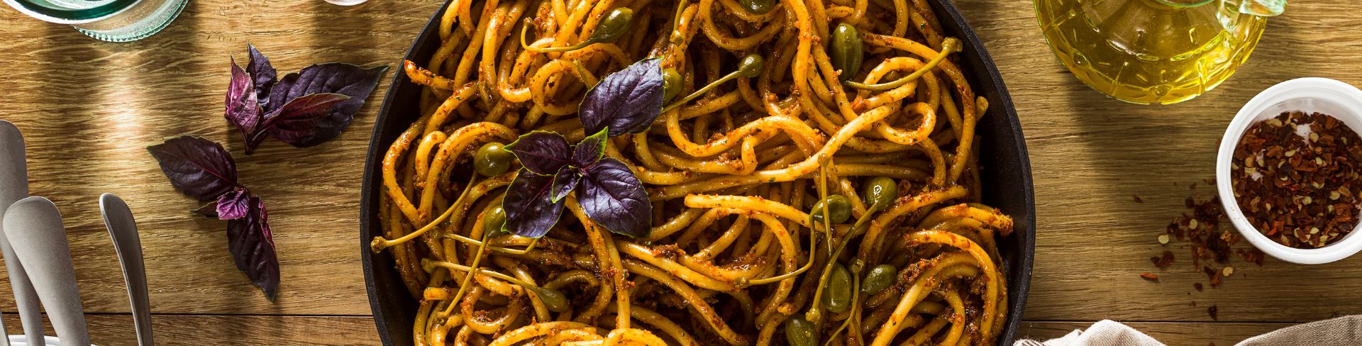 Top-down view of a bowl of bucatini pasta with pesto and caper berries.