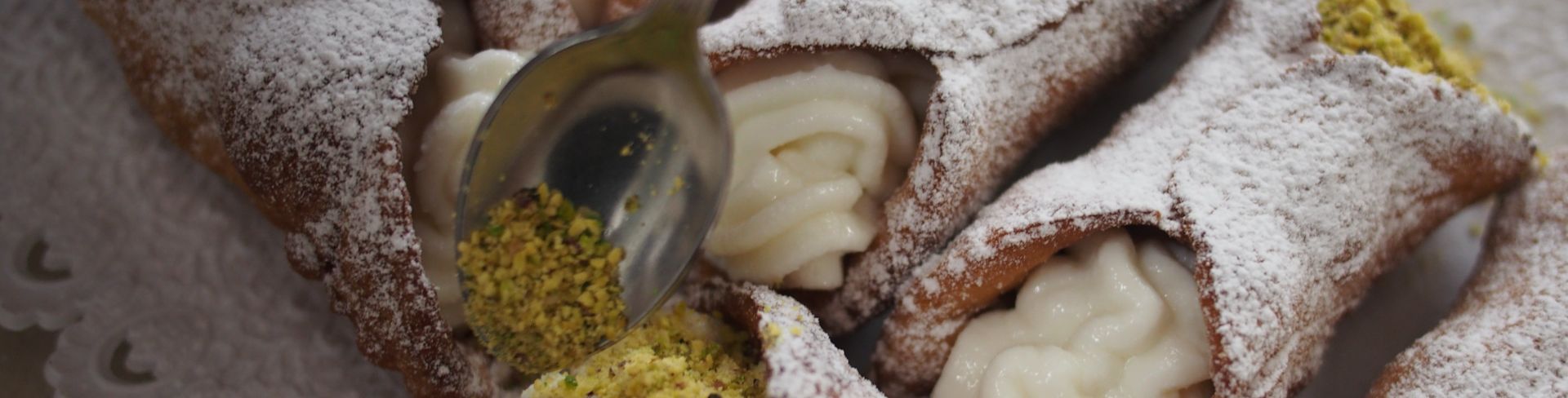 Someone spoons crumbled pistachios onto the end of a filled cannoli.