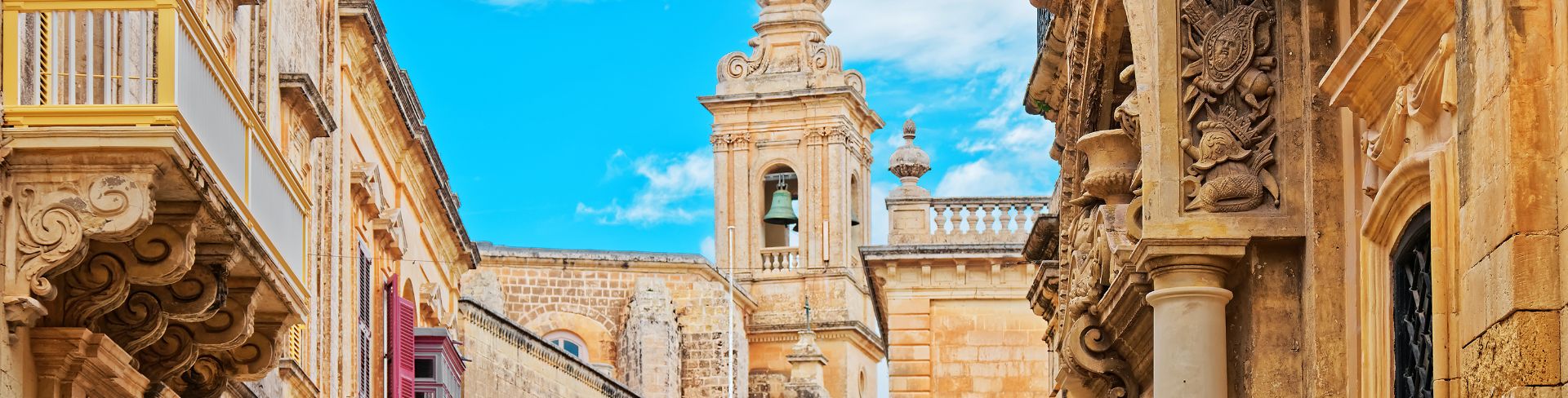 Looking between lavish stone buildings of Malta at the bell tower of Palazzo Santa Sofia.